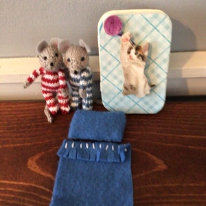 May include: Three gray knitted mice wearing pajamas, a blue blanket, and a small cardboard box with a blue and white checkered pattern and a picture of a white kitten with a purple ball of yarn.