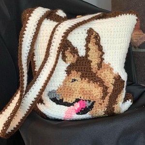 May include: A white crocheted bag with a brown strap and a brown and tan crocheted German Shepherd dog design. The dog is looking to the left and has a pink tongue sticking out.