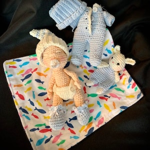 May include: A crocheted baby doll with a pacifier, wearing a diaper and blue booties, laying on a white blanket with a colorful fish pattern. A blue crocheted outfit and a crocheted bunny are also on the blanket.