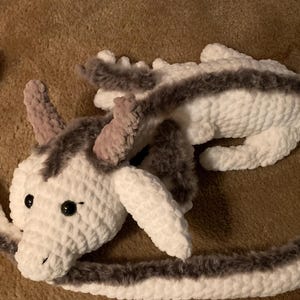 May include: A white crocheted dragon with a long, gray and white tail. The dragon has a fluffy texture and black eyes.