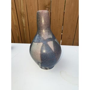 Op de afbeelding: Een keramische vaas met een bolvormig lichaam en een lange, slanke hals. De vaas is beschilderd met een mix van kleuren, waaronder blauw, grijs en paars. Het ontwerp heeft geometrische vormen. De vaas is ongeveer 25 cm hoog.