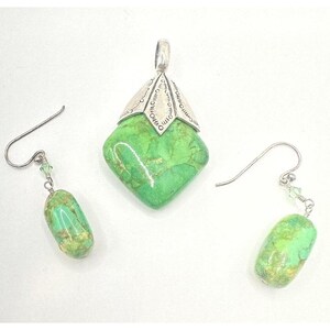 Può includere: Un set di gioielli composto da un ciondolo in pietra verde a forma di diamante con una chiusura in metallo color argento e due orecchini pendenti abbinati con pietre verdi cilindriche sospese a ganci color argento con piccole perline.