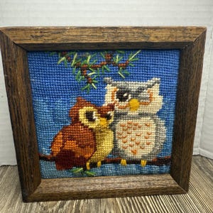 May include: A framed needlepoint featuring two owls perched on a branch. The owls are rendered in shades of brown, yellow, gray, and white against a blue background. The frame is made of dark wood, adding a rustic touch to the artwork.