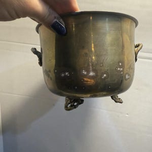 Op de afbeelding: Een aangetaste messing plantenbak met drie sierlijke voetjes en twee handvatten aan de zijkant. De kleine, ronde container heeft een licht uitlopende rand en een verweerde uitstraling, wat duidt op leeftijd en gebruik. De plantenbak wordt door een hand vastgehouden.