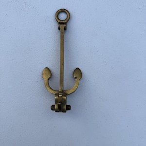 May include: A brass anchor-shaped decorative object. The anchor has a ring at the top, a long central shaft, and curved arms with rounded ends. The anchor is made of brass and has a vintage appearance.