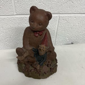 May include: A brown teddy bear figurine with a red bow tie, sitting next to a smiling gnome wearing a red hat and black coat. The base is a textured brown with small details.