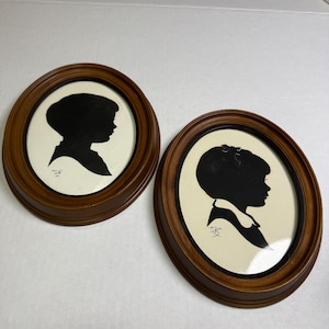 May include: Two oval wall frames made of medium-toned wood, each containing a black silhouette portrait of a child against a cream-colored background. The silhouettes show side profiles of children, one with a bow in their hair, and both feature a small signature at the bottom.