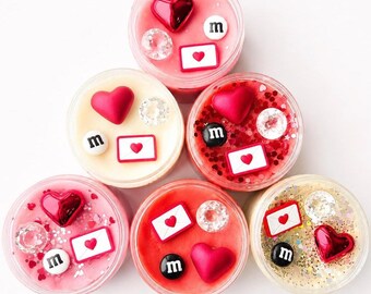 May include: Six circular containers filled with colorful slime, each topped with decorative charms including red hearts, white envelopes with red hearts, clear faceted beads, and small white or black circular tokens featuring the letter m.