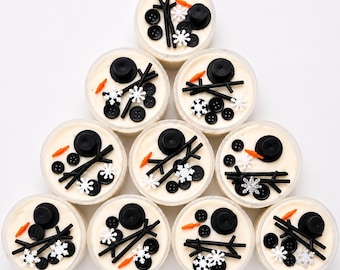 May include: Ten clear circular containers filled with cream-colored substance, each decorated with black buttons, orange carrot-shaped pieces, black stick-like accents, and white snowflake embellishments arranged to resemble snowman faces.