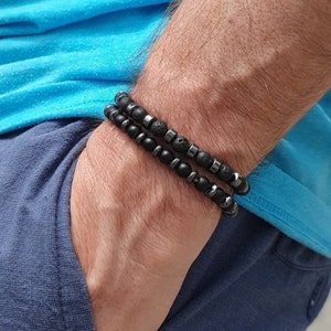Könnte beinhalten: Zwei schwarze Perlenarmbänder, die auf einem Handgelenk übereinander liegen. Ein Armband besteht aus schwarzen Lavasteinen und das andere aus schwarzen Hämatitperlen.