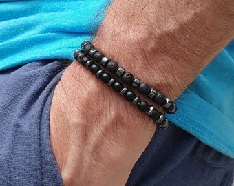 Könnte beinhalten: Zwei schwarze Perlenarmbänder, die auf einem Handgelenk übereinander liegen. Ein Armband besteht aus schwarzen Lavasteinen und das andere aus schwarzen Hämatitperlen.