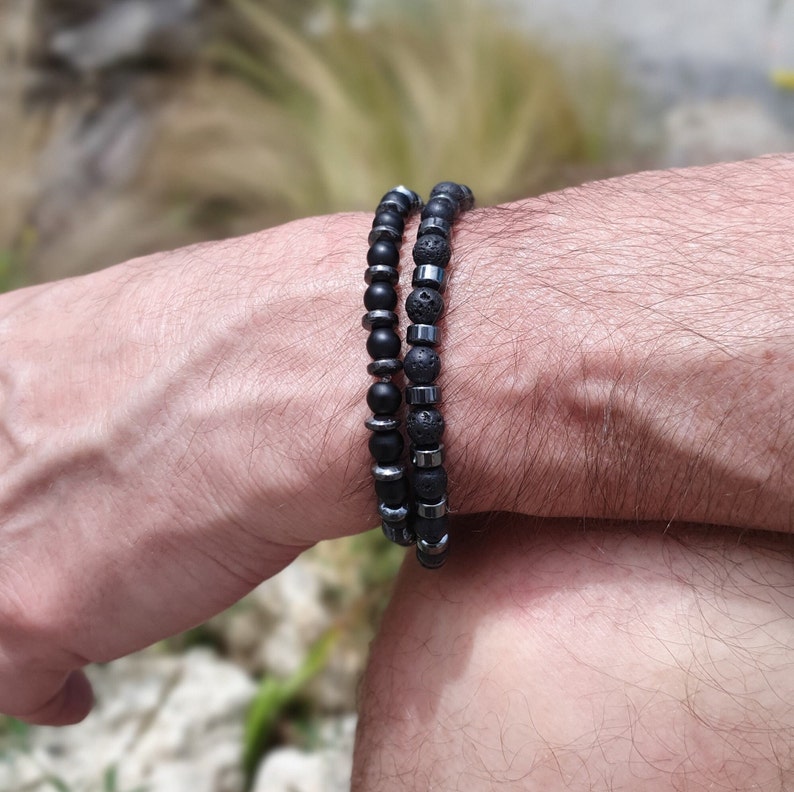 Peut inclure: Deux bracelets en perles sur le poignet d'une personne. Un bracelet est fait de perles de lave noire et l'autre de perles d'onyx noir.