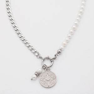 Puede incluir: Un collar de cadena de plata con un dije de plata que presenta un diseño intrincado. La cadena está conectada a una hebra de perlas blancas.