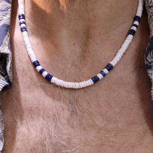 Könnte beinhalten: Eine weiße und blaue Muschelkette mit einem schlichten Design. Die Kette besteht aus kleinen, runden Muscheln, die auf einer Schnur aufgefädelt sind.