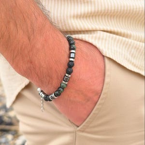 May include: A beaded bracelet featuring a combination of dark green, black, and silver beads. The bracelet is composed of round and square beads, with a chain and clasp closure. The bracelet is worn on a wrist.