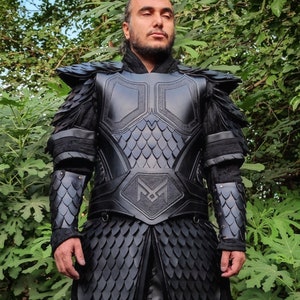 Puede incluir: Una armadura de cuero negro con un patr&oacute;n de escamas y una gran letra "M" estilizada en el pecho. La armadura tiene un cuello alto y cubre los brazos y los hombros.