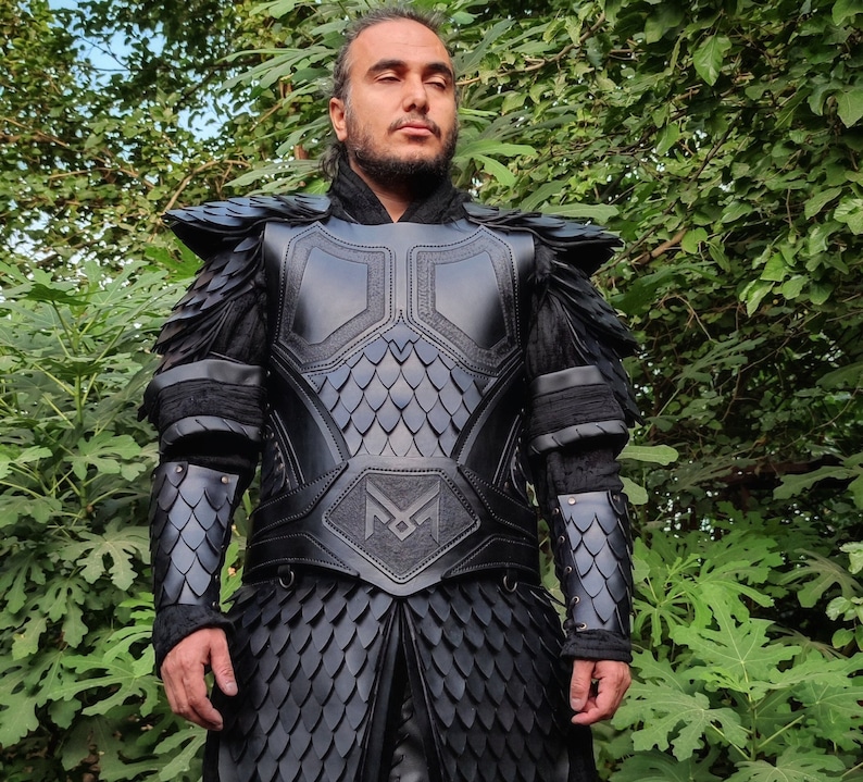 Puede incluir: Una armadura de cuero negro con un patr&oacute;n de escamas y una gran letra "M" estilizada en el pecho. La armadura tiene un cuello alto y cubre los brazos y los hombros.