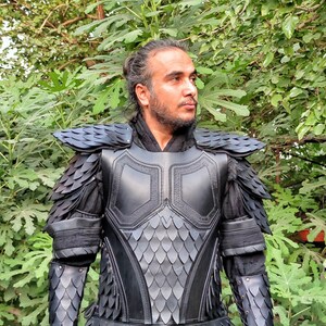 Puede incluir: Una armadura de cuero negro con un patr&oacute;n de escamas. La armadura tiene una pieza de pecho, hombreras y protectores de brazos. La armadura la lleva puesta una persona con el pelo largo y oscuro.
