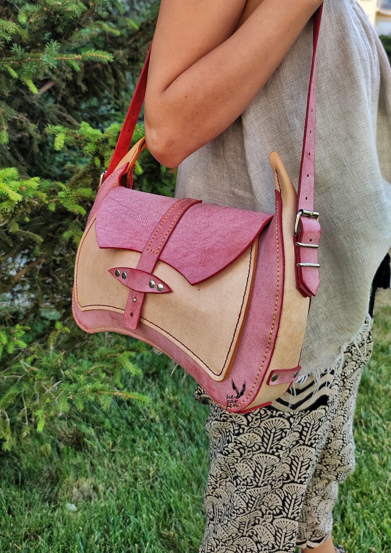 Op de afbeelding: Een roze en beige leren schoudertas met een klep sluiting en een zilveren gesp. De tas heeft een gestikt ontwerp en een klein logo op de bodem.