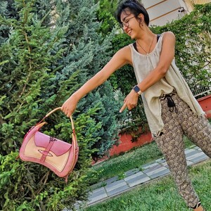 Op de afbeelding: Een vrouw in een wit tanktop en een broek met patroon houdt een roze en beige leren handtas voor een groene struik.