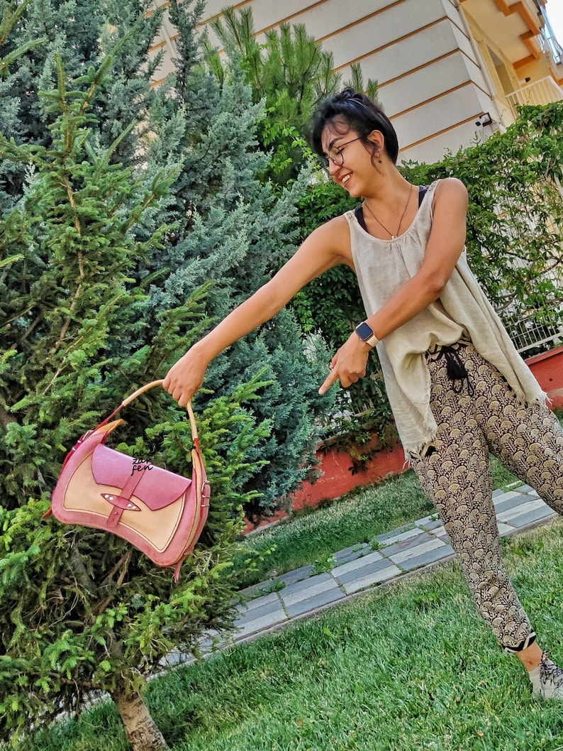 Op de afbeelding: Een vrouw in een wit tanktop en een broek met patroon houdt een roze en beige leren handtas voor een groene struik.
