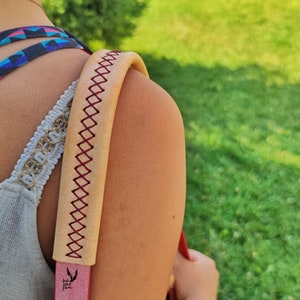 Op de afbeelding: Een tan lederen schouderband met rode stiksels en een klein zwart logo met de tekst "he for she".
