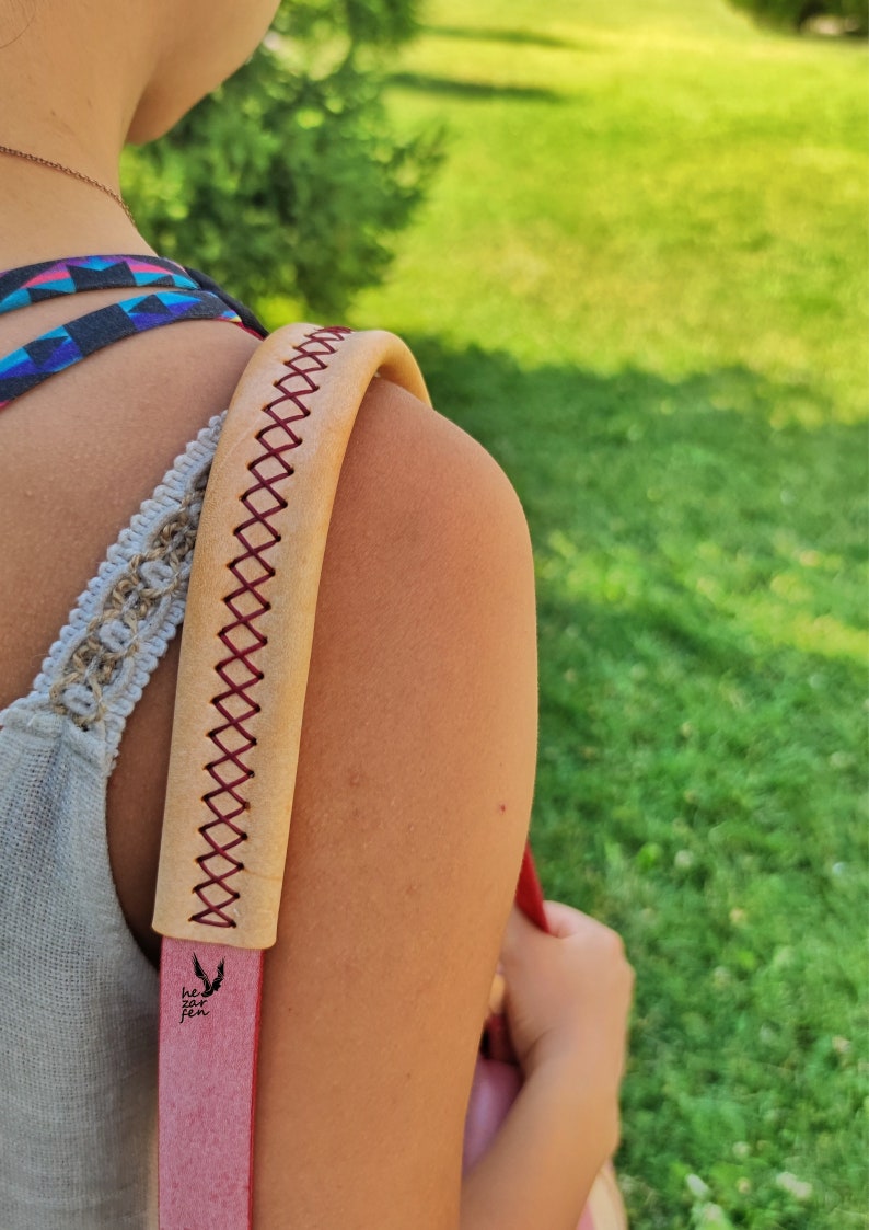 Op de afbeelding: Een tan lederen schouderband met rode stiksels en een klein zwart logo met de tekst "he for she".