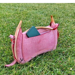 Op de afbeelding: Een roze leren handtas met een klep sluiting en een klein vakje aan de voorkant. Een groene telefoon is in het vakje gestopt. De tas heeft een bruine leren band en een klein metalen gesp. De tas ligt op een bed van groen gras.