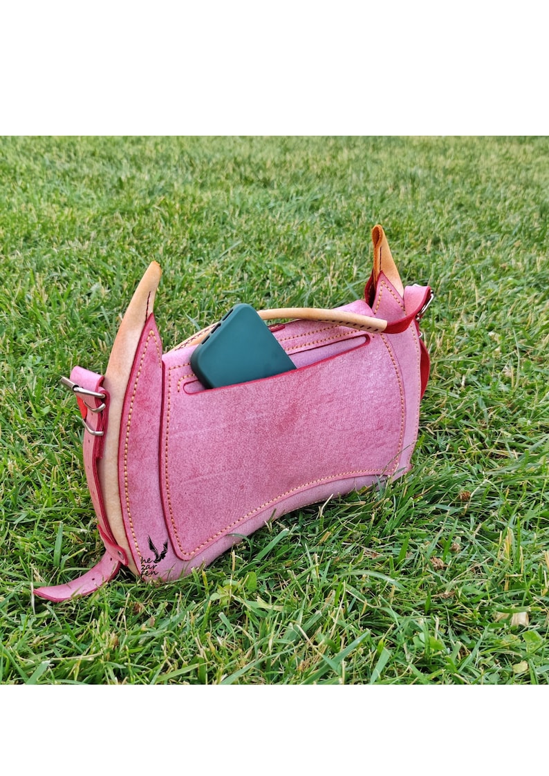 Op de afbeelding: Een roze leren handtas met een klep sluiting en een klein vakje aan de voorkant. Een groene telefoon is in het vakje gestopt. De tas heeft een bruine leren band en een klein metalen gesp. De tas ligt op een bed van groen gras.