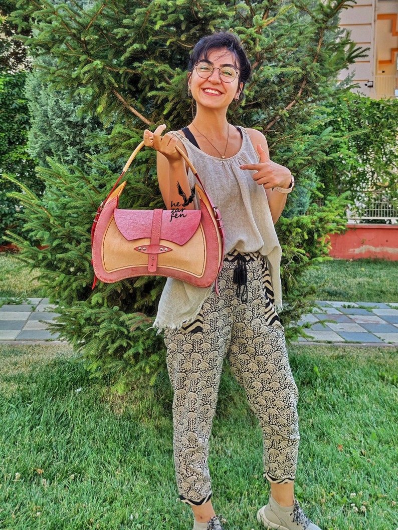 Op de afbeelding: Een vrouw houdt een roze en beige leren handtas vast met een houten handvat. De tas heeft een klep met een metalen gesp en een klein vakje aan de voorkant. Op de tas staat de tekst "hezar fen".