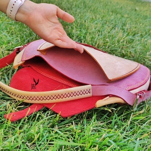 Op de afbeelding: Een roze leren tas met een beige klep en een rode voering. De tas heeft gele stiksels en een klein logo met de tekst "he zur fen".