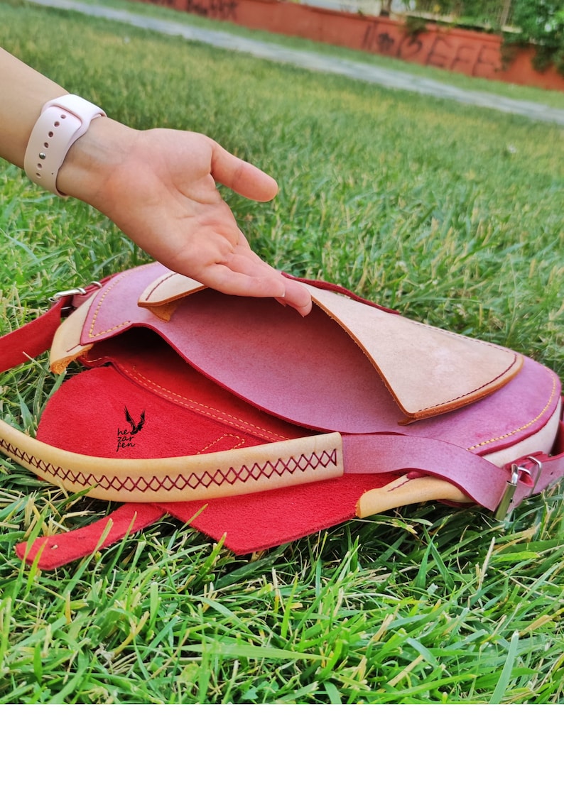Op de afbeelding: Een roze leren tas met een beige klep en een rode voering. De tas heeft gele stiksels en een klein logo met de tekst "he zur fen".