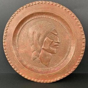 Puede incluir: Un plato decorativo redondo de color cobre con un perfil en relieve de una persona. El plato tiene un borde con cuentas y un diseño en relieve detallado. El perfil está centrado dentro de un marco circular.