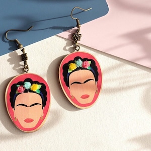 May include: A pair of wooden earrings featuring a portrait of Frida Kahlo with a floral crown. The earrings are painted in shades of pink, blue, green, and yellow. The earrings are attached to bronze-coloured hooks.