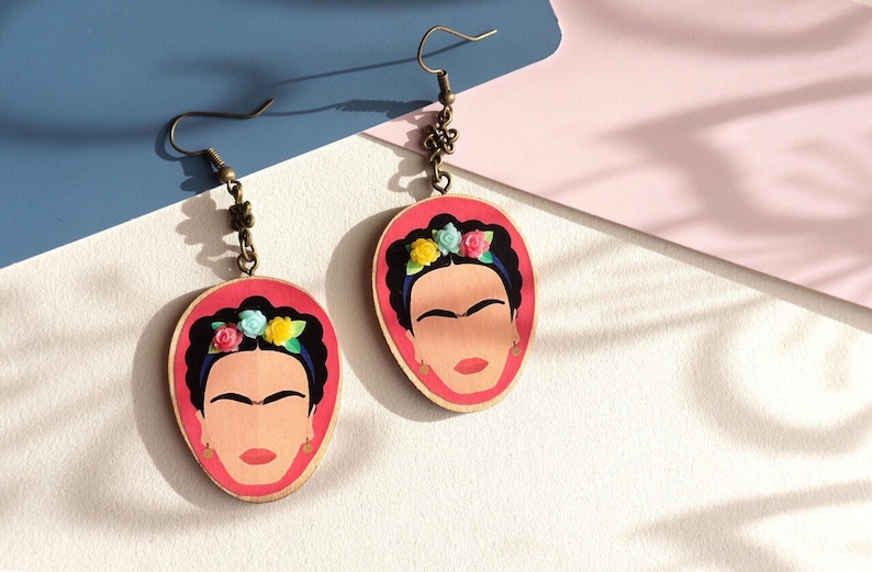 May include: A pair of wooden earrings featuring a portrait of Frida Kahlo with a floral crown. The earrings are painted in shades of pink, blue, green, and yellow. The earrings are attached to bronze-coloured hooks.
