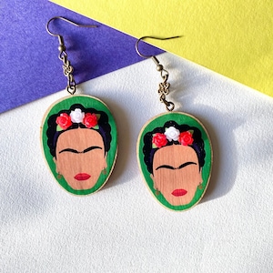 May include: A pair of green wooden earrings featuring a painted portrait of Frida Kahlo, with red and white flowers in her hair.
