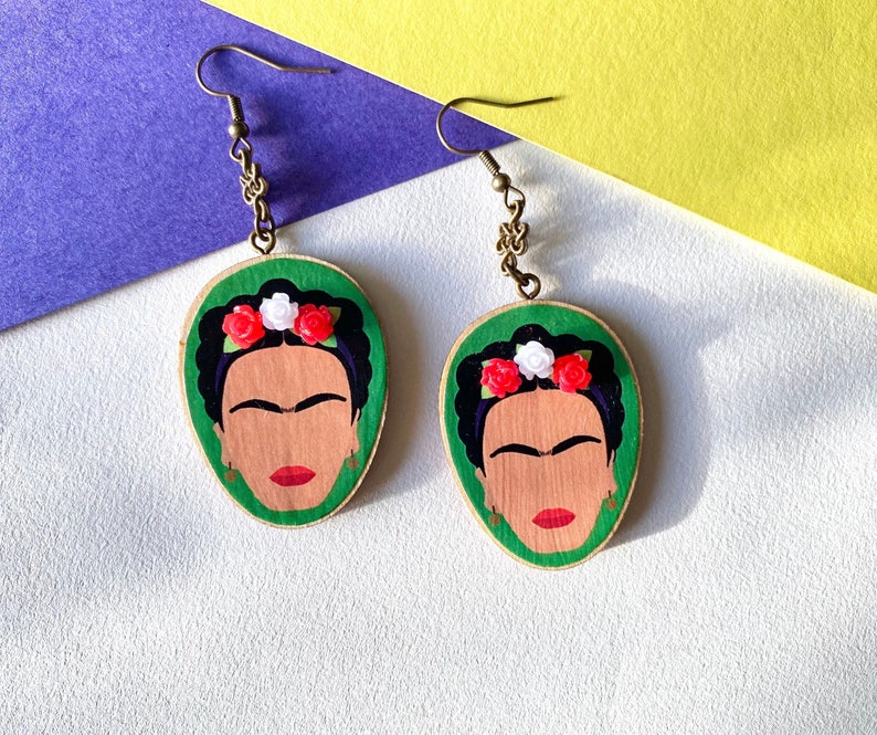 May include: A pair of green wooden earrings with a painted portrait of Frida Kahlo, featuring a red and white flower crown.