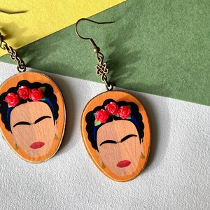 May include: A pair of wooden earrings featuring a painted portrait of Frida Kahlo with red roses in her hair. The earrings have a gold-toned hook.