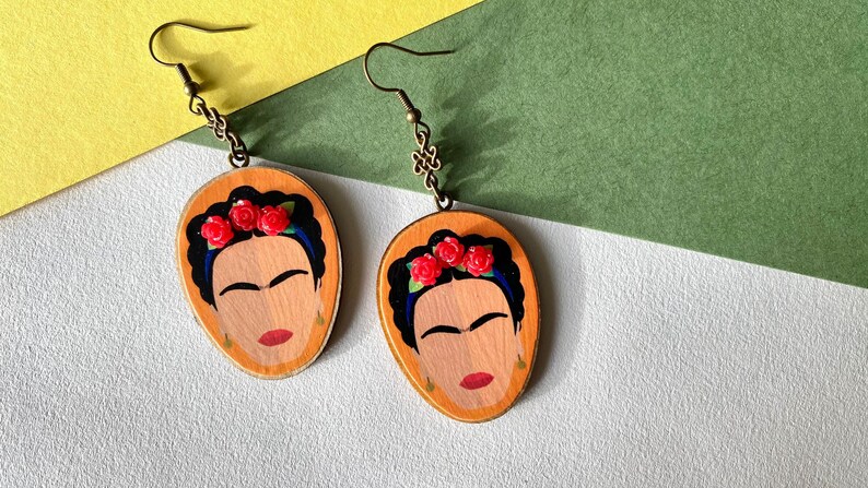 May include: A pair of wooden earrings featuring a painted portrait of Frida Kahlo with red roses in her hair. The earrings have a gold-toned hook.