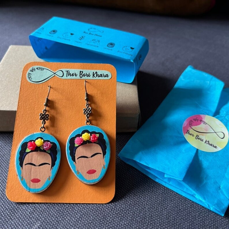 May include: A pair of earrings featuring a painted portrait of Frida Kahlo on a blue background. The earrings are made of wood and have a bronze hook. The earrings are packaged in a blue box with the text "Thor Bori Khara" on it.