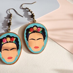 May include: A pair of earrings featuring a blue background with a portrait of Frida Kahlo. The earrings are made of wood and have a bronze-coloured hook.