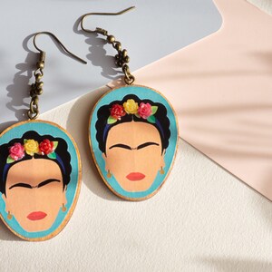 May include: A pair of dangle earrings featuring a blue background with a portrait of Frida Kahlo. The portrait is surrounded by a wreath of colourful flowers. The earrings have a bronze chain and hook.