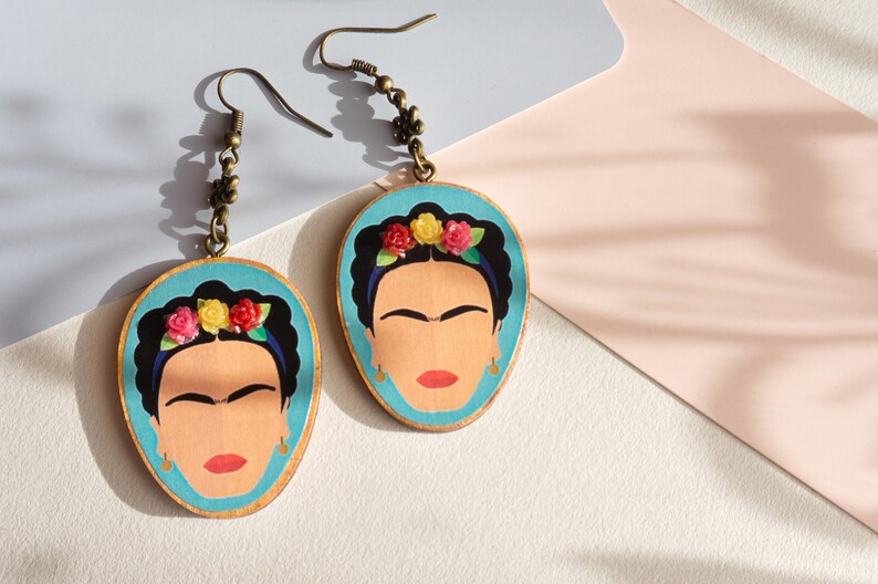 May include: A pair of dangle earrings featuring a blue background with a portrait of Frida Kahlo. The portrait is surrounded by a wreath of colourful flowers. The earrings have a bronze chain and hook.