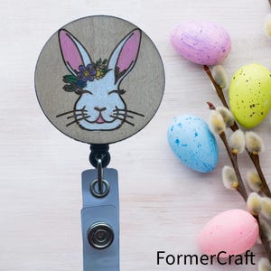 May include: A wooden badge reel featuring a white bunny with pink ears and a flower crown. The bunny is smiling and has a light blue nose. The badge reel is surrounded by pastel coloured Easter eggs and a branch with willow buds.