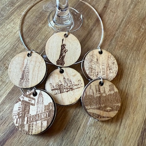 May include: Six wooden wine glass charms with different images of New York City landmarks, including the Statue of Liberty, the Empire State Building, the Brooklyn Bridge, and Central Park. The charms are attached to a silver ring.
