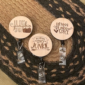 May include: Three wooden badge reels with fall-themed designs. The reels feature the phrases "Lil Miss Pumpkin Spice" with pumpkins and hearts, and "Pumpkin Spice Junkie" with an arrow. The reels are on a textured brown and black surface.