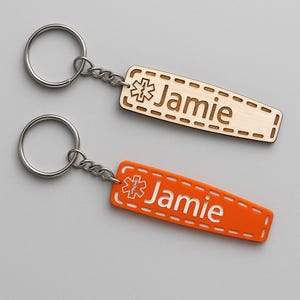 Może przedstawiać: Dwa breloki do kluczy ze srebrnymi kółkami i łańcuszkami. Jeden jest z naturalnego drewna z imieniem "Jamie" i symbolem medycznym. Drugi jest pomarańczowy z tym samym imieniem i symbolem. Oba mają szytą obwódkę.