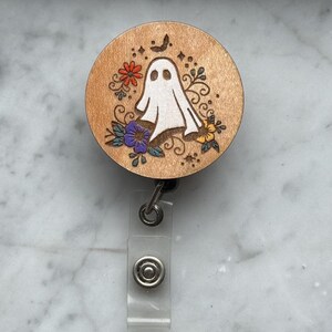Op de afbeelding: Een houten badge reel met een Halloween thema. De ronde reel heeft een wit spookontwerp omringd door kleurrijke bloemen en een kleine vleermuis. De reel heeft een doorzichtige plastic band en een metalen clip.
