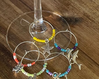 Könnte beinhalten: Eine Auswahl an Weinglas-Anhängern mit silbernen Ringen, die mit bunten Perlen und Anhängern zum Thema Meer verziert sind. Die Charms umfassen einen Seestern, Schildkröten, einen Delfin und einen Anker, ideal für Partys.