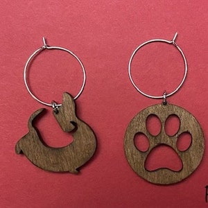 May include: A pair of wooden earrings with silver hoops. One earring features a silhouette of a dachshund dog, while the other earring features a paw print.